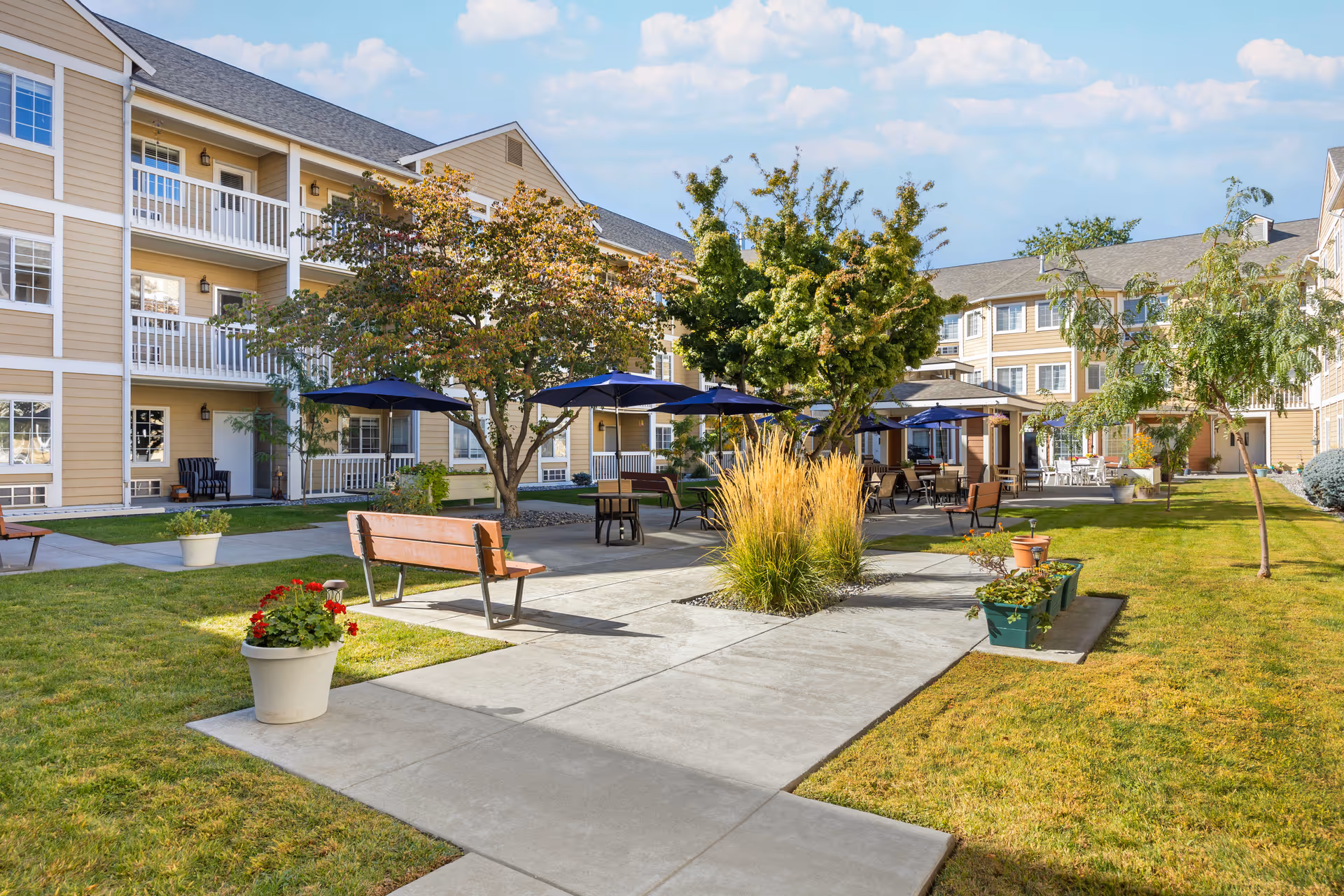Courtyard of a multi-story senior living building with benches, umbrellas, potted plants, and walkways.