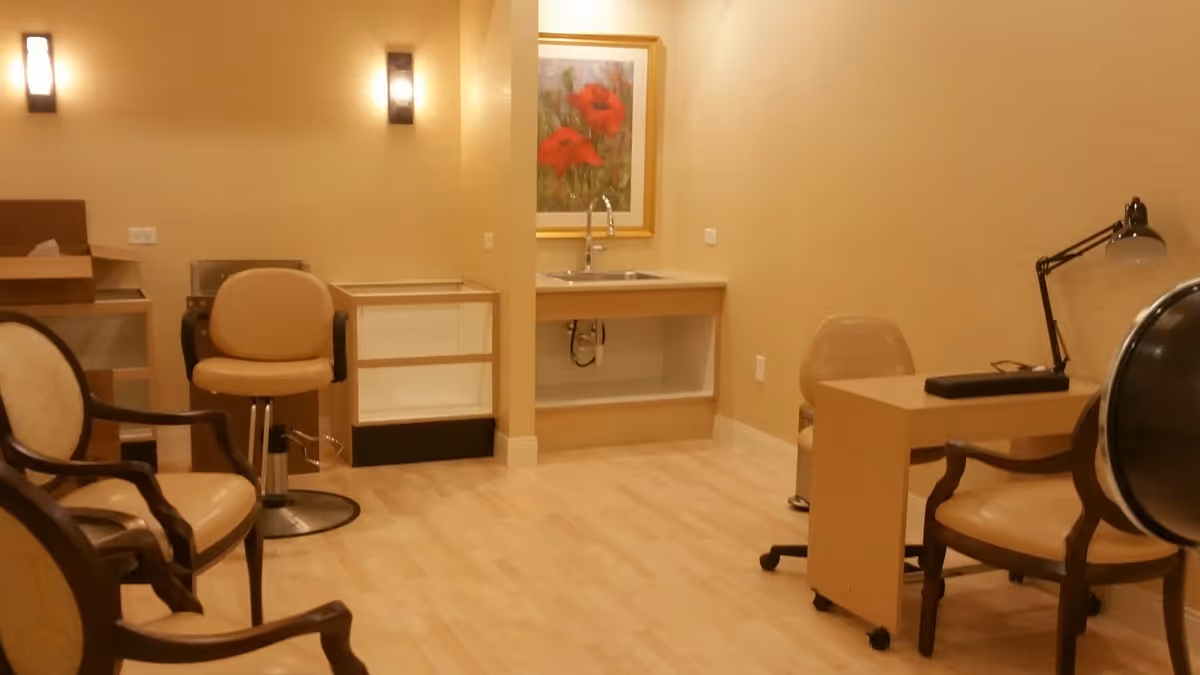 Small interior room with salon chairs, a desk, a sink under a framed floral painting, and soft lighting.
