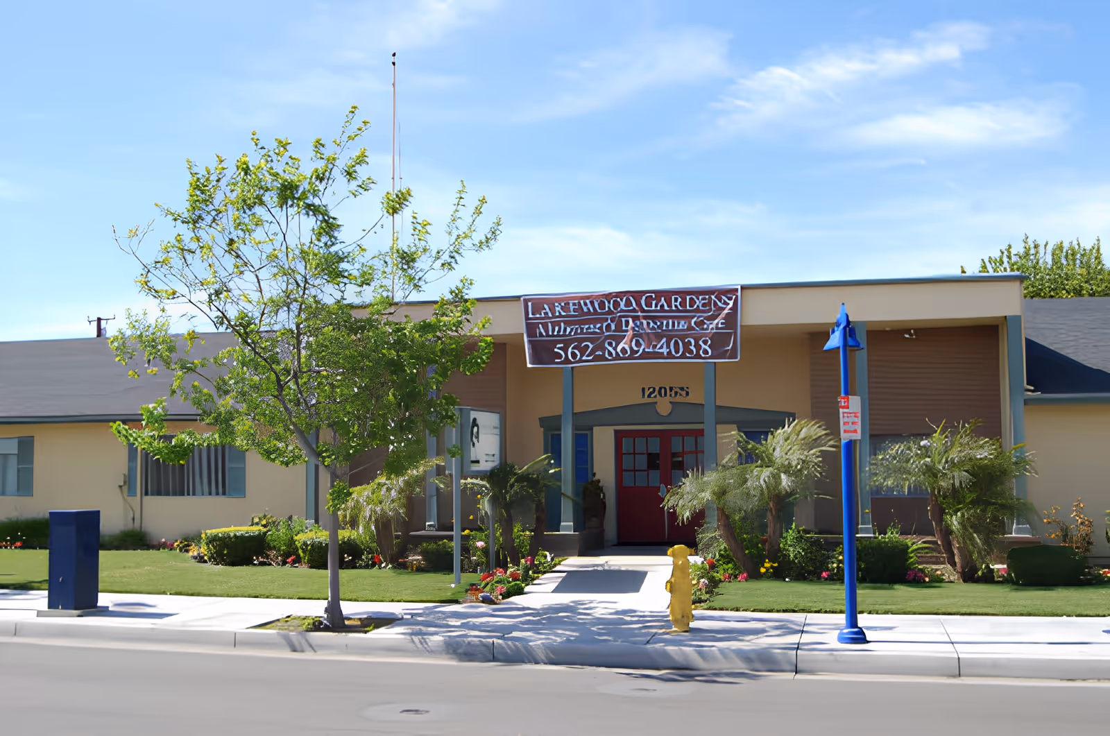 Front exterior view of Lakewood Gardens Alzheimer's & Dementia Care facility with a beige building, red entrance door, green landscaping, a tree, and a blue street lamp on the sidewalk.