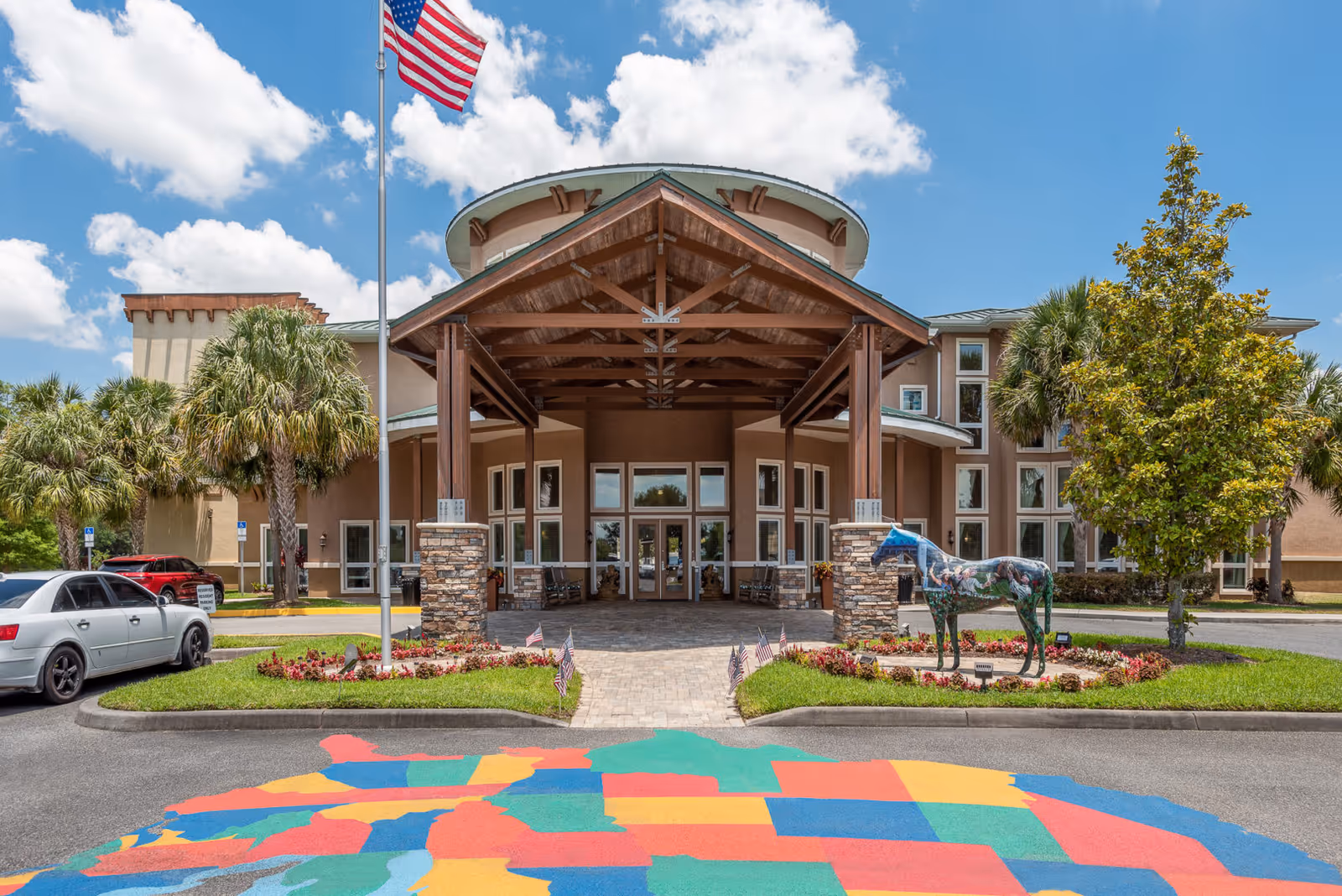 Front exterior view of Brentwood at Fore Ranch facility with a covered entrance supported by wooden beams and stone pillars. An American flag flies on a flagpole in front, and a colorful painted map of the United States is visible on the driveway. There are palm trees, a decorative horse statue, and parked cars near the entrance under a partly cloudy blue sky.