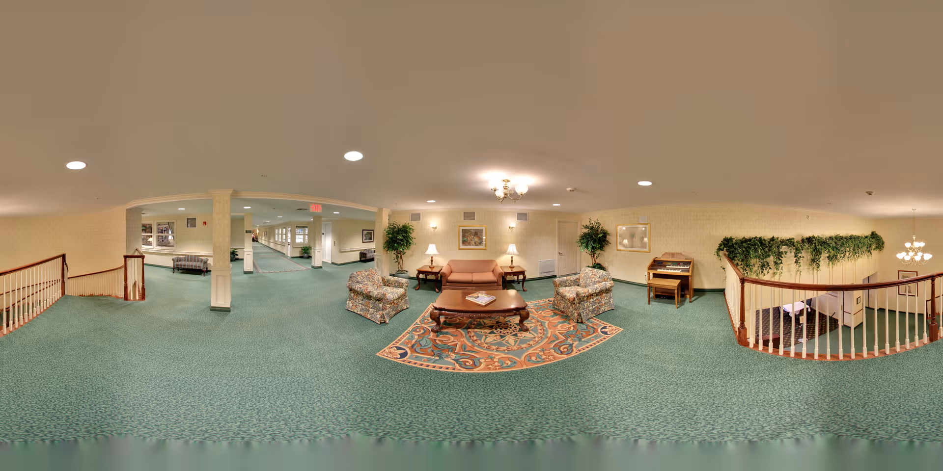 A spacious senior living facility common area with green carpet, floral armchairs, a pink sofa, a wooden coffee table on a patterned rug, two side tables with lamps, framed artwork on the walls, potted plants, and a piano. The area is well-lit with ceiling lights and a chandelier, and there is a staircase with wooden railing leading downstairs.