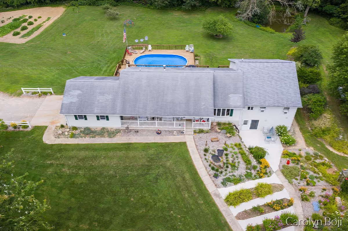 Aerial view of a single-story building with a gray roof surrounded by green lawns and landscaped gardens. There is a small swimming pool with a deck and chairs behind the building. A flagpole with an American flag is near the pool. The property includes a garden area with plants and flowers, and a paved walkway leading to the building entrance.