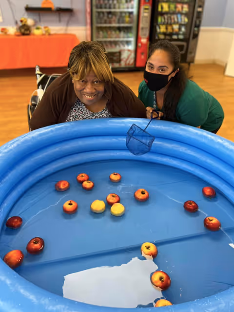 Two women smiling and leaning over a blue inflatable pool filled with water and floating apples, one woman holding a small net. The setting appears to be indoors with vending machines and a table with a red tablecloth in the background.