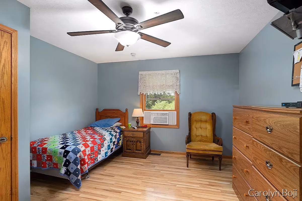 A simple bedroom with a single bed covered by a colorful quilt, wooden nightstand, mustard armchair, dresser, ceiling fan, and a window with an air conditioner.