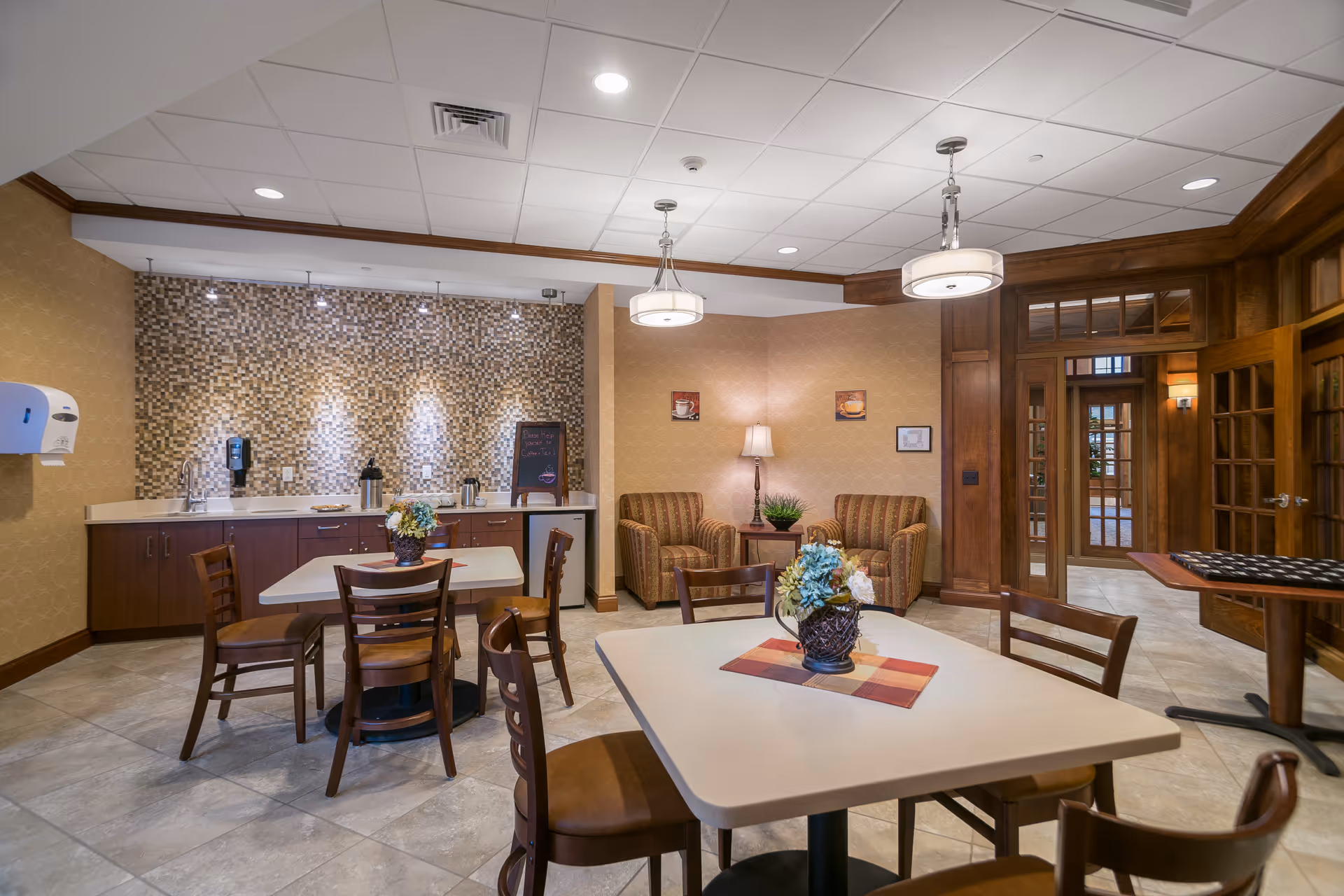 Bright communal dining and lounge area with tables, chairs, a kitchenette with a mosaic backsplash and upholstered seating.
