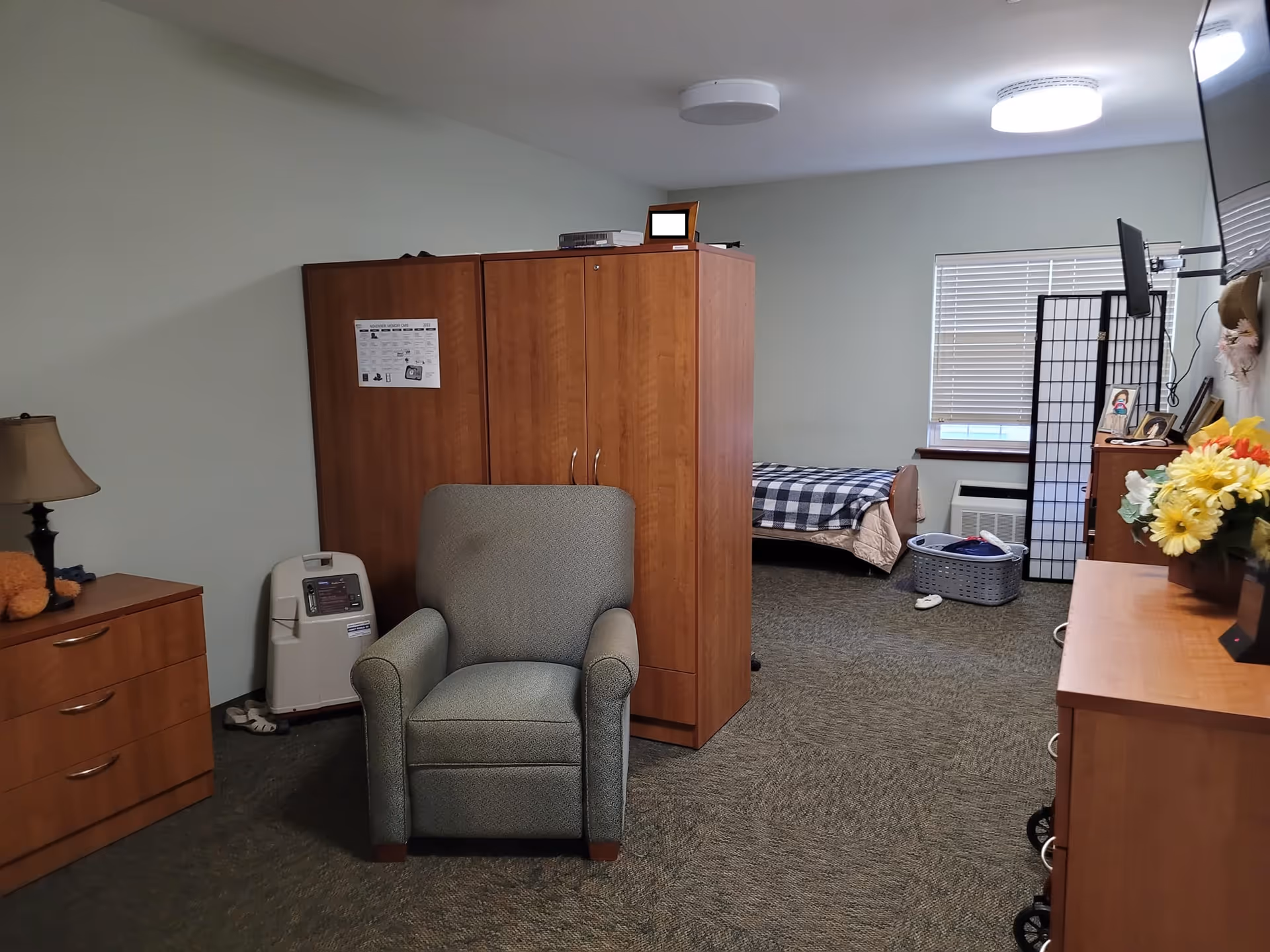 A senior living facility bedroom with a single bed covered with a checkered blanket near a window with blinds. The room has wooden furniture including a wardrobe, a dresser with a lamp and flowers, and a TV mounted on the wall. There is a comfortable armchair in the center and a laundry basket on the floor.