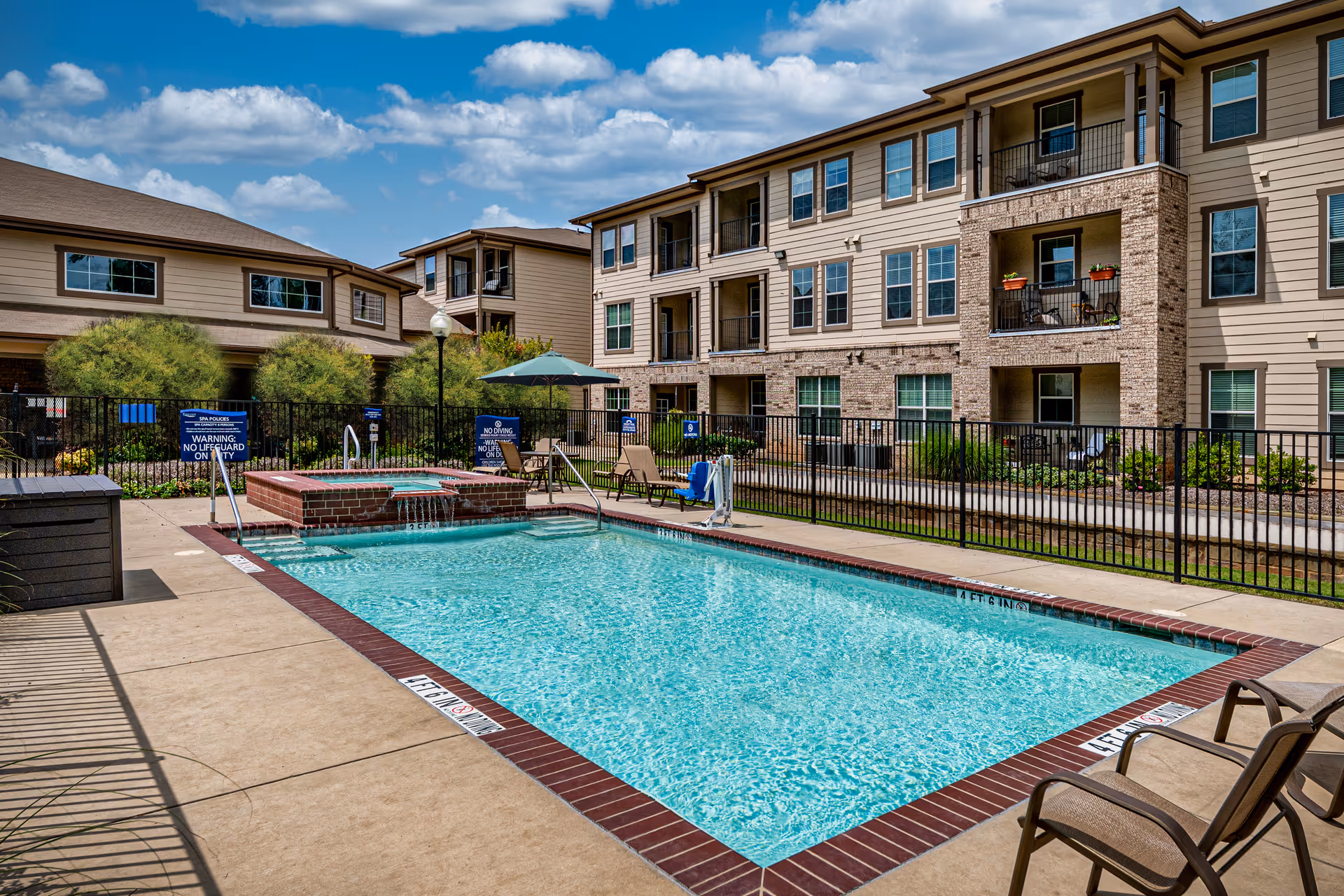 Outdoor swimming pool area at Parkview on Hollybrook with clear blue water, surrounded by a concrete deck and several lounge chairs. There is a hot tub adjacent to the pool with water flowing into the pool. The pool area is fenced, and behind it are multi-story residential buildings with balconies and windows. The sky is partly cloudy with blue patches.