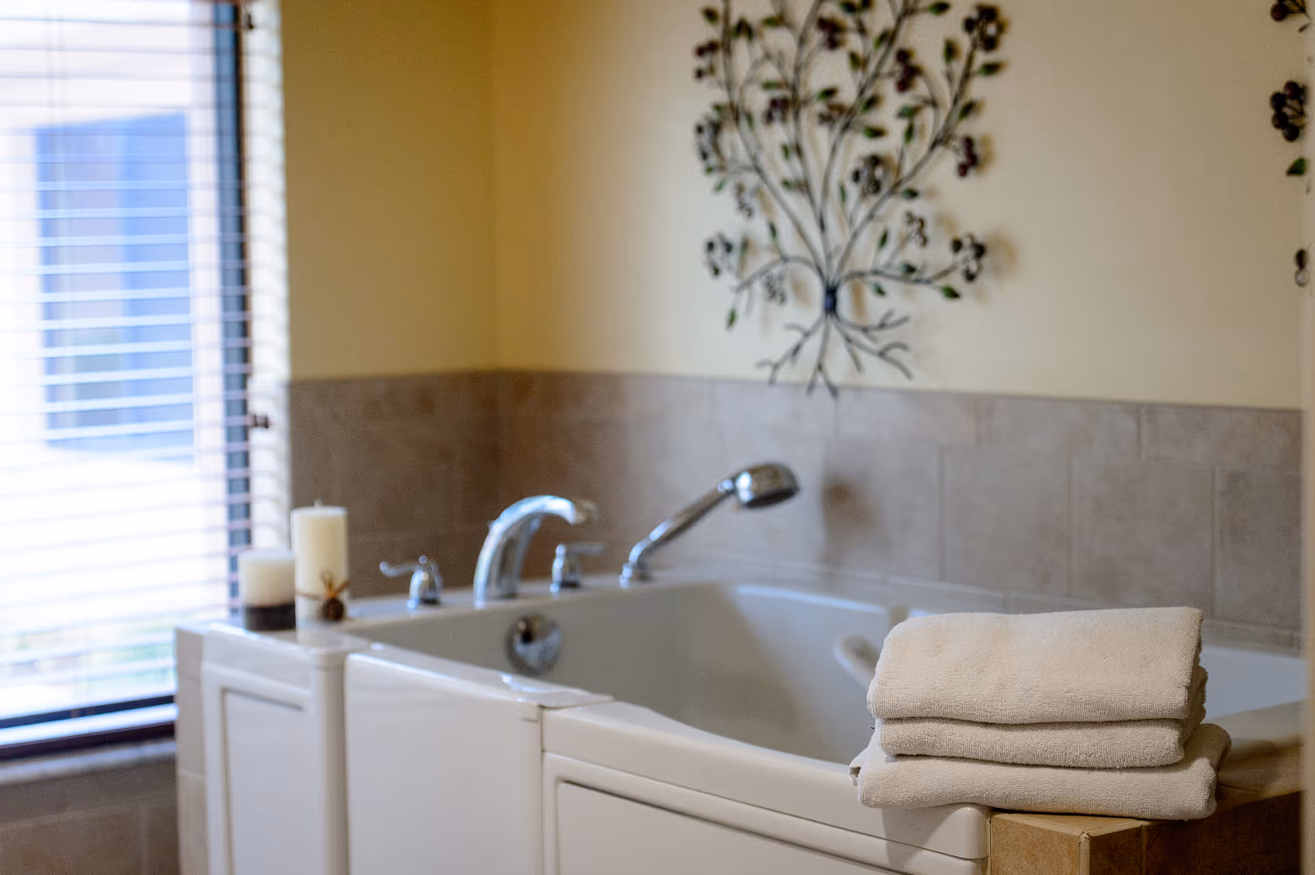 A bathroom featuring a white bathtub with chrome fixtures, a stack of folded white towels on the edge, two candles on the corner, a window with blinds allowing natural light, and a decorative metal wall art above the tub.