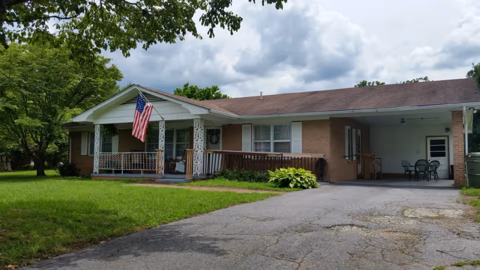 Single-story brick house with a front porch, American flag, carport and driveway in a grassy yard.