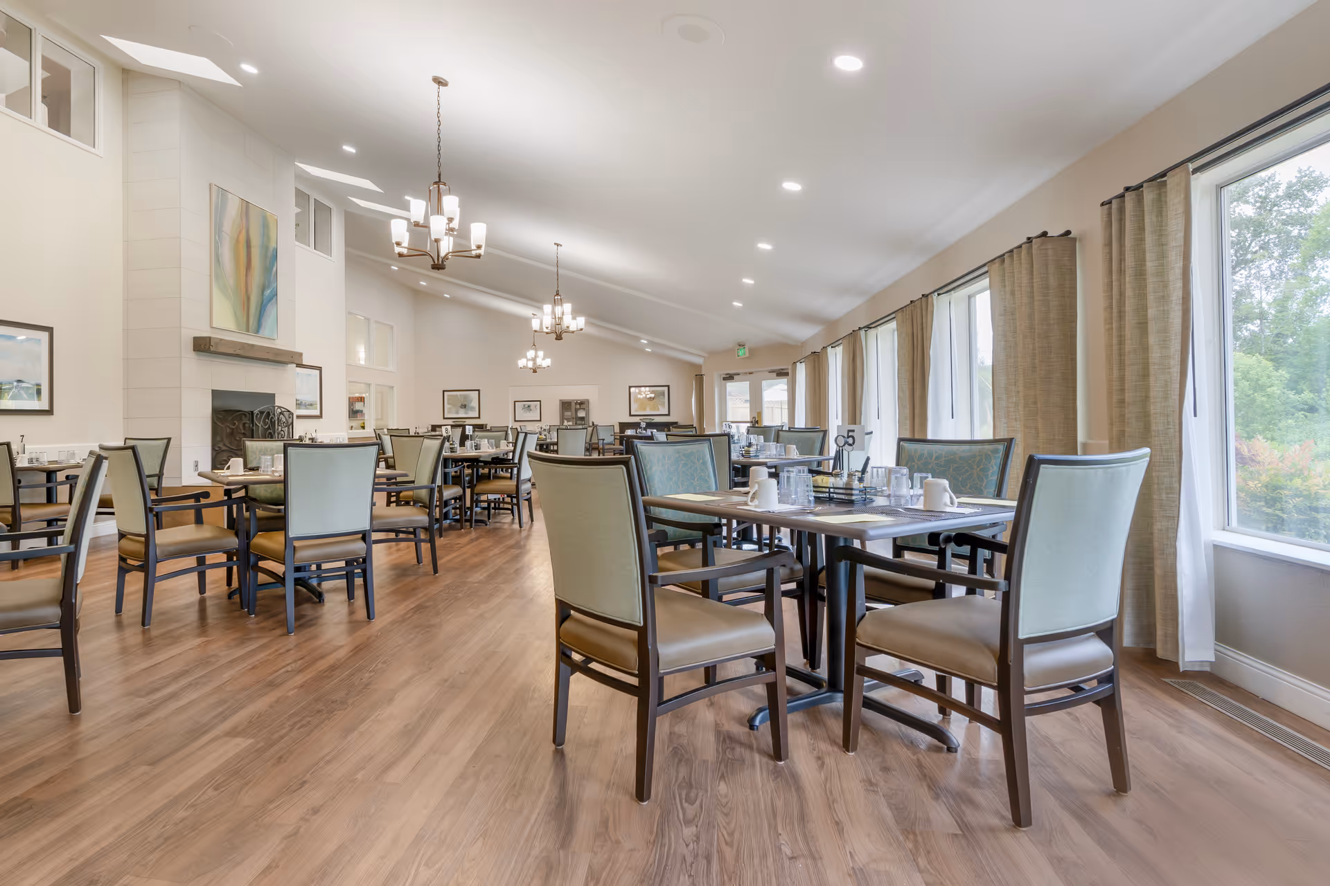 A spacious dining room with multiple tables and chairs arranged neatly. The room features large windows with beige curtains allowing natural light to brighten the space. There are chandeliers hanging from the ceiling and framed artwork on the walls. The floor is wooden, and the tables are set with cups, glasses, and condiments.
