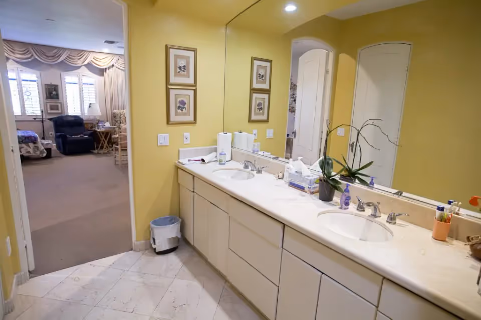 Bathroom vanity with double sinks and a large mirror, yellow walls, and a view into an adjoining bedroom/living area.