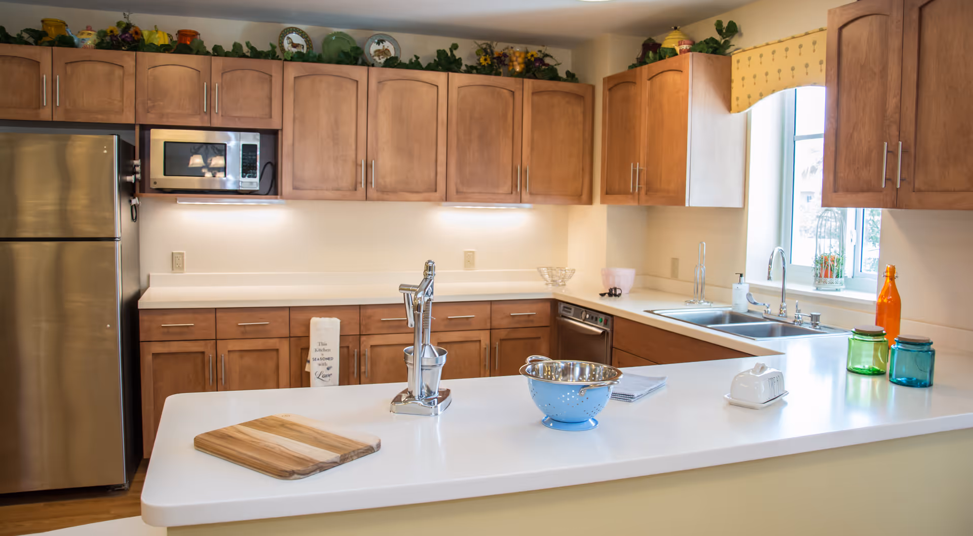 A bright kitchen with wooden cabinets, a stainless steel refrigerator, microwave, and dishwasher. The kitchen features a white countertop island with a cutting board, a metal colander, a citrus juicer, and colorful glass jars. There is a window with a yellow valance above the sink, and decorative plates and plants are displayed above the cabinets.