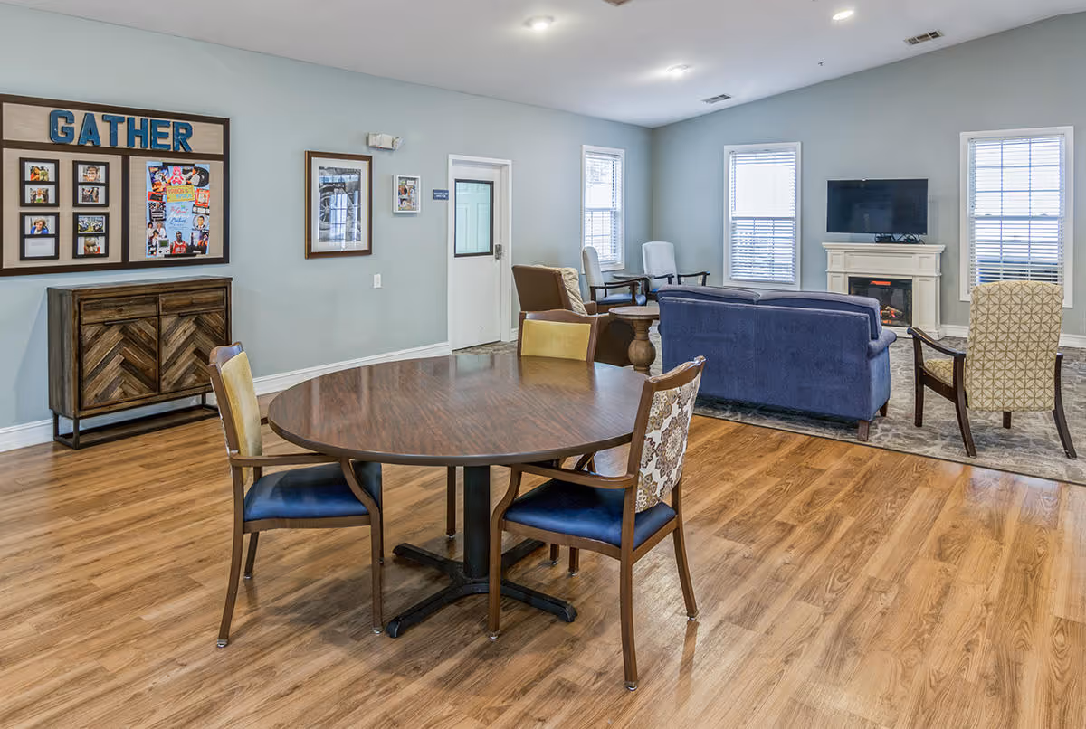 A spacious common area in Dalton Square featuring a round wooden table with four chairs in the foreground. In the background, there is a seating area with a blue couch, two armchairs, a fireplace with a TV mounted above it, and three windows with blinds. The walls are light blue, and there is a wooden cabinet with a photo collage frame labeled 'GATHER' on the left wall.