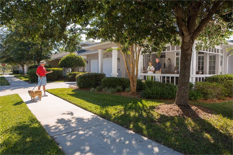 A man wearing a red shirt and hat is walking a dog on a leash along a sidewalk in a residential area with well-maintained lawns and trees. Two people are standing on the porch of a light-colored house with white columns and large windows, engaged in conversation.