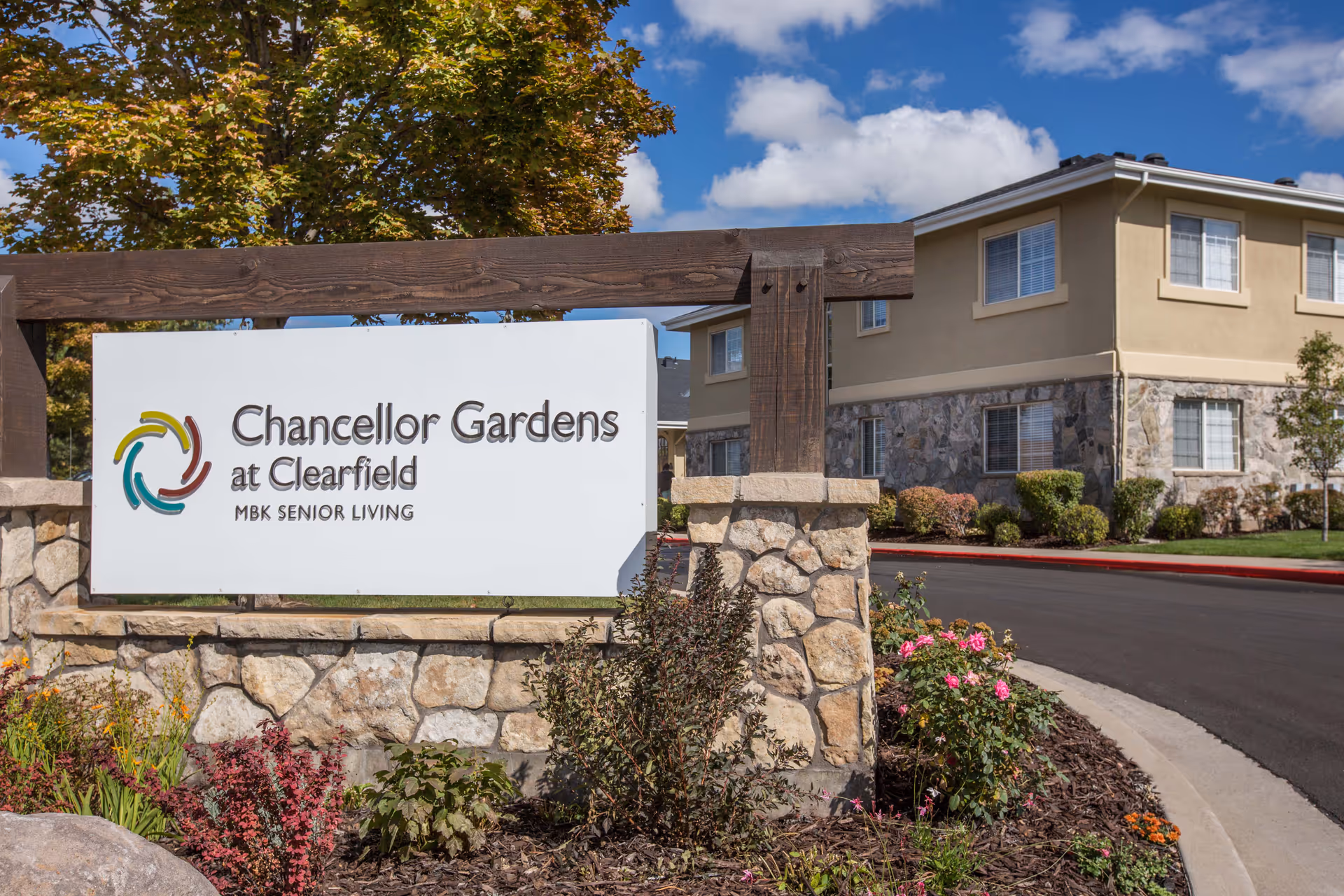 Outdoor view of the entrance sign for Chancellor Gardens at Clearfield, MBK Senior Living, with a stone and wood structure surrounded by landscaped plants and flowers, and a beige building with stone accents in the background under a blue sky with clouds.