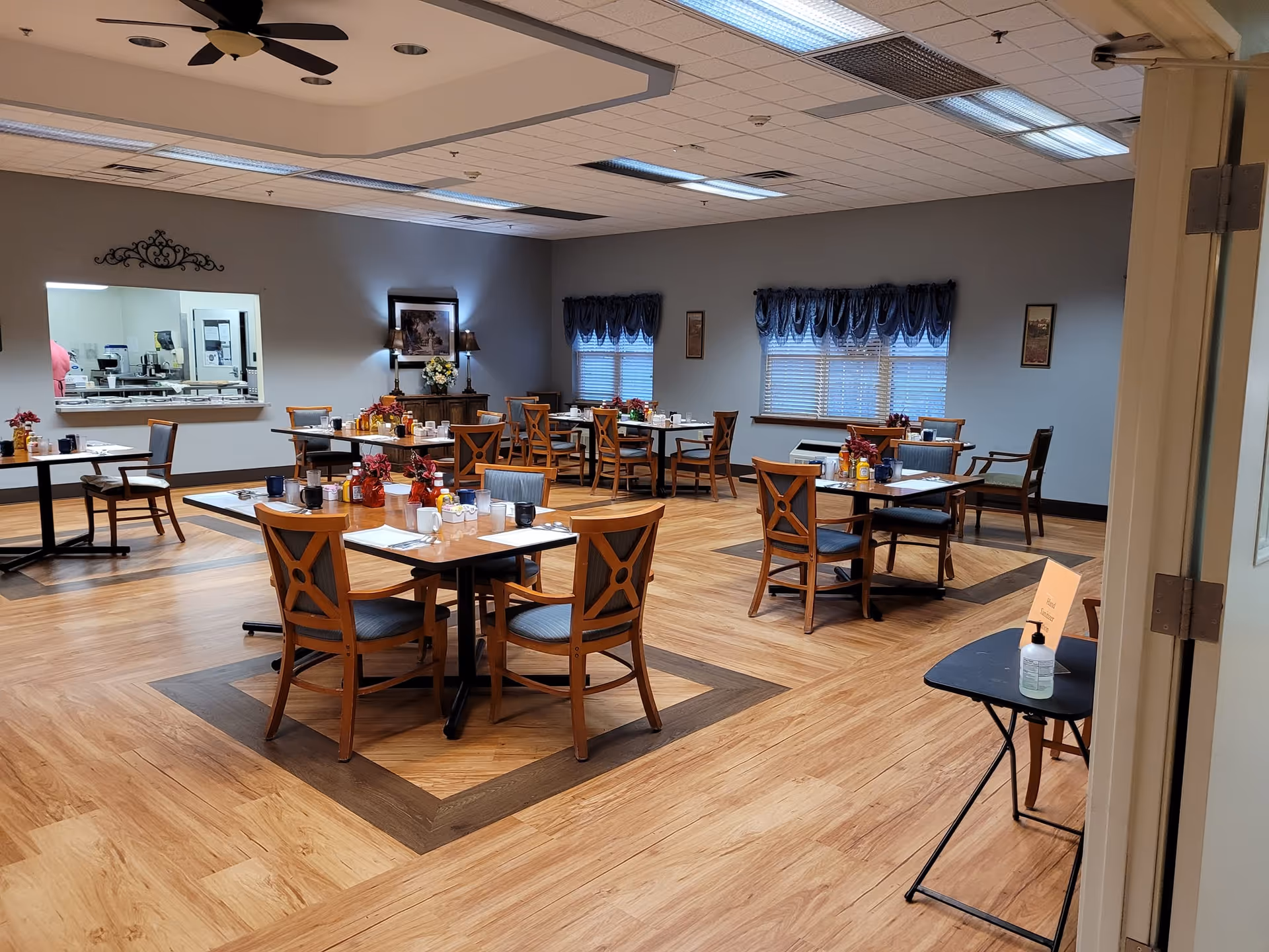 A dining room in The Gardens of Green Ridge facility with several wooden tables and chairs arranged neatly. Each table is set with placemats, cups, and condiments. The room has wood-patterned flooring with darker wood inlays around the tables, gray walls, and windows with blue valances. A serving window to the kitchen is visible on the left wall, and a small table with hand sanitizer and a sign is near the entrance on the right.