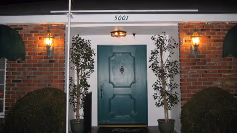 Front entrance of a building with a green door flanked by two potted plants and two wall-mounted lantern-style lights on either side. The building has a brick exterior and the number 5001 is displayed above the door.