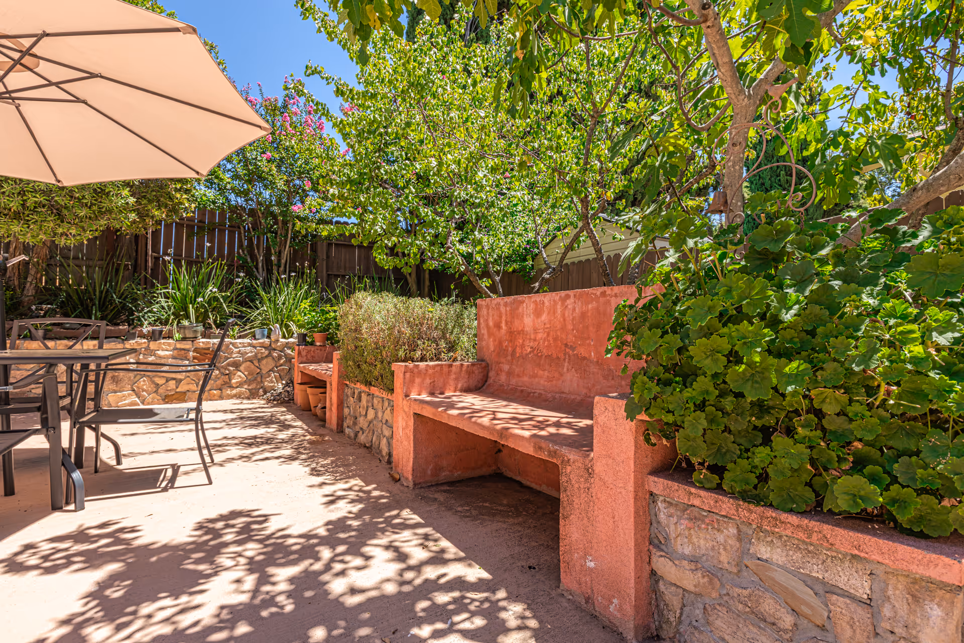 Sunlit outdoor patio with a stone bench, potted plants, a dining table and umbrella, surrounded by trees and a wooden fence.