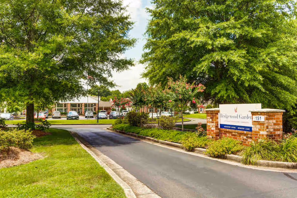 Entrance driveway to Bridgewood Gardens senior living facility with a brick sign displaying the facility name and services offered, surrounded by green trees and landscaped bushes, with parked cars and the building visible in the background.