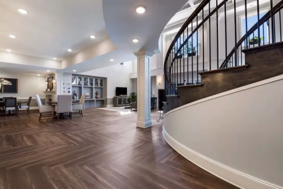 Spacious interior of a senior living facility featuring a curved staircase with dark wooden steps and black railing, a seating area with a TV and built-in shelves, and a dining area with tables and chairs. The room has wood-patterned flooring, white walls, and recessed ceiling lights.