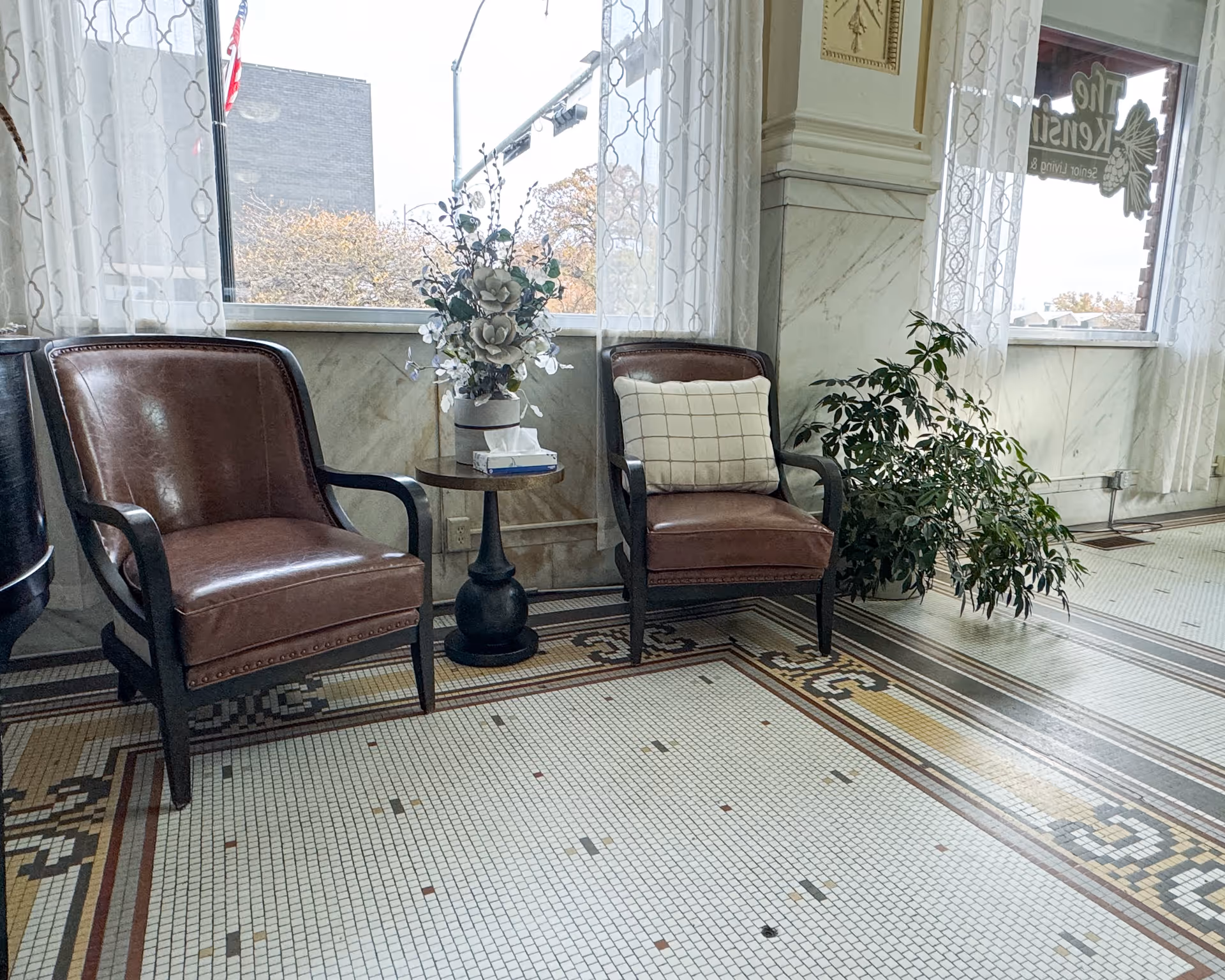 Two brown leather armchairs with wooden frames are placed on a decorative tiled floor next to a small round table holding a vase with white flowers and a box of tissues. Sheer white curtains cover the windows behind the chairs, and a green potted plant is positioned near the right chair. The window on the right has a sign that reads 'The Kensington'.