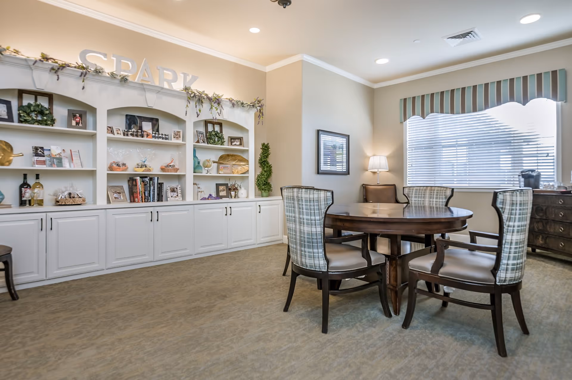 A well-lit room with a round wooden table surrounded by four upholstered chairs with plaid backs. Behind the table is a large window with blinds and a striped valance. To the left, there is a built-in white shelving unit decorated with plants, books, framed photos, and decorative items, with the word 'SPARK' displayed on top. The room has beige walls and carpeted flooring.