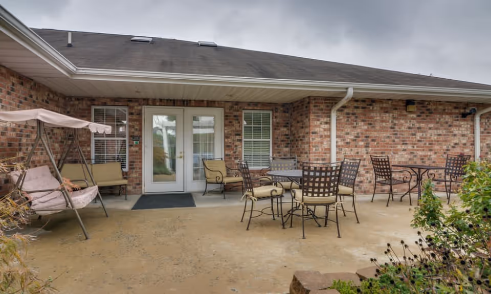 Outdoor patio area at Auburn Creek Senior Living with brick walls, a covered porch, cushioned metal chairs around tables, and a cushioned swing seat. There are two white-framed glass doors and windows with white blinds.