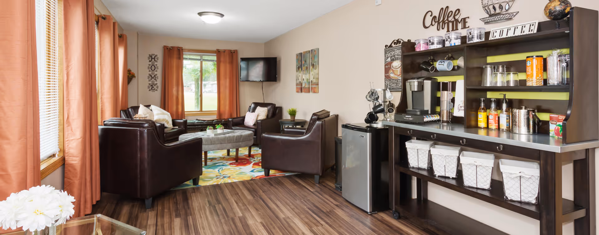 A cozy living room area with dark brown leather chairs arranged around a colorful rug and a central ottoman. The room has large windows with orange curtains letting in natural light. On the right side, there is a coffee station with various coffee supplies, a mini fridge, and decorative items on the wall including the words 'Coffee Time'.