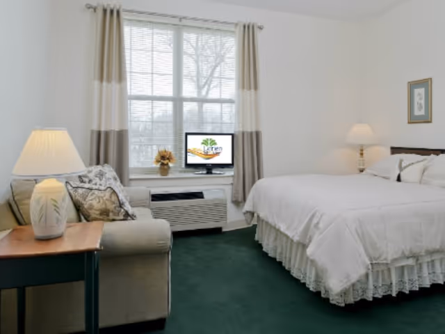 A cozy bedroom with a large bed covered in white bedding, a beige sofa with patterned pillows, a wooden side table with a lamp, a window with beige and white curtains, and a small TV on the windowsill displaying the Lorien logo.