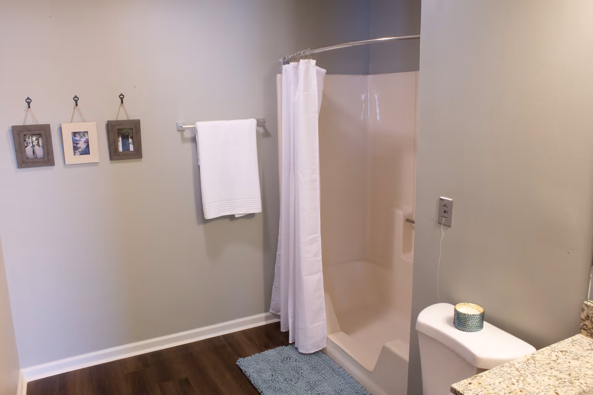 A bathroom with a white shower curtain partially closed around a shower stall, a white towel hanging on a towel rack, three small framed pictures on the wall, a blue bath mat on the wooden floor, and a toilet with a candle on top next to a granite countertop.