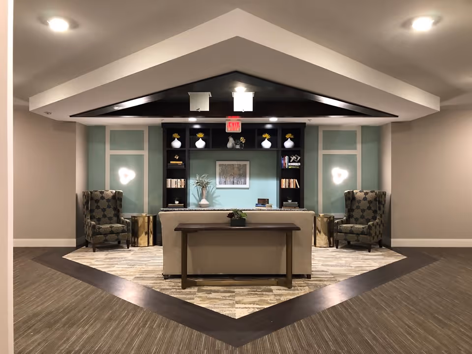 A cozy and modern common area in a senior living facility featuring a beige sofa with a wooden console table behind it, two patterned armchairs on either side, gold side tables, a built-in black bookshelf with decorative vases and books, a framed artwork on a light blue accent wall, and soft overhead lighting.