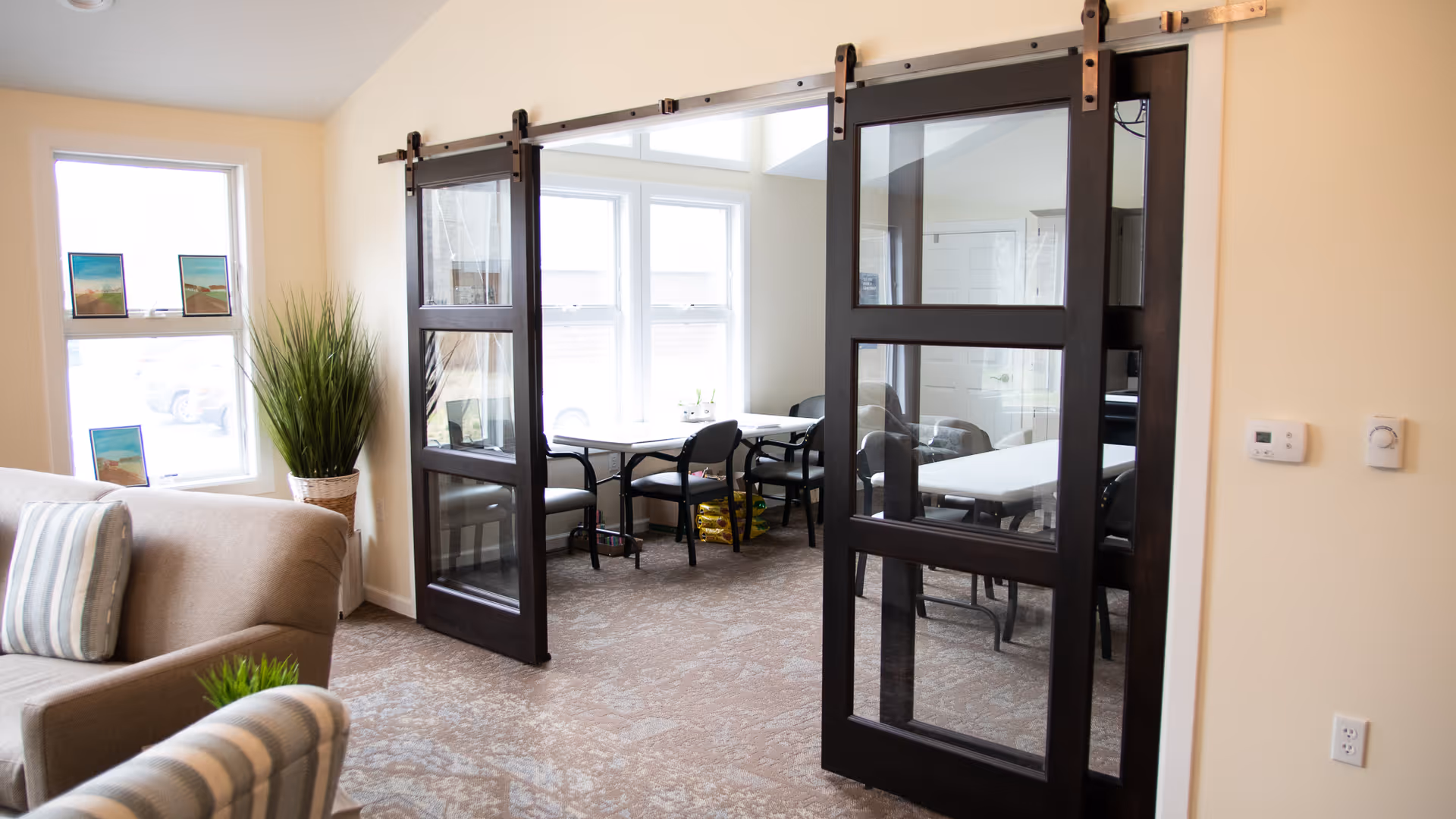 A bright interior room with beige walls and carpeted floor featuring two dark wooden sliding glass doors. Beyond the doors is a small dining or meeting area with white tables and black chairs near large windows letting in natural light. In the foreground, there is a beige armchair with striped cushions and a potted plant near a window.