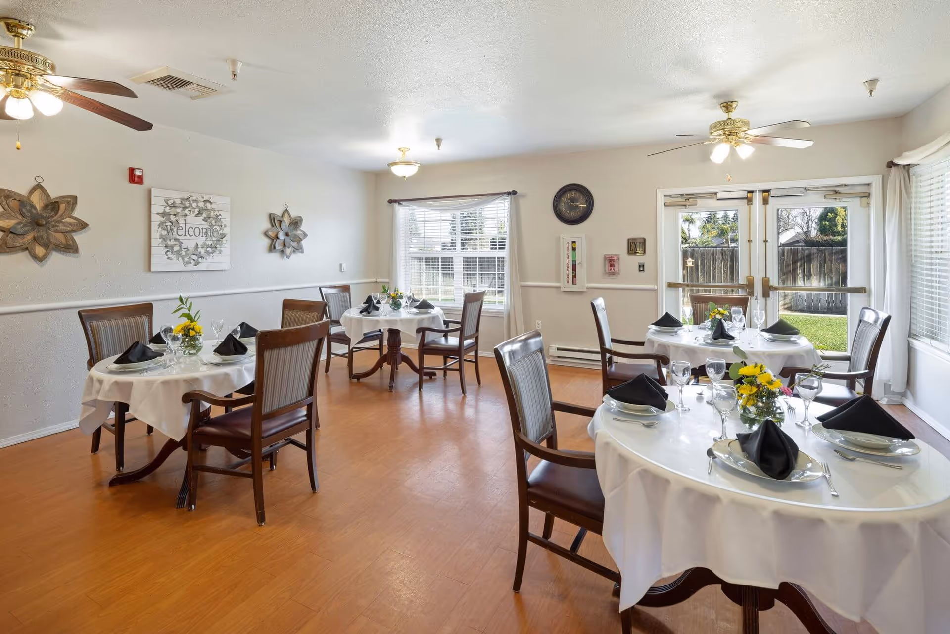 Bright dining room with round tables set with white tablecloths, black napkins and floral centerpieces, wooden chairs, windows and double doors to a yard.