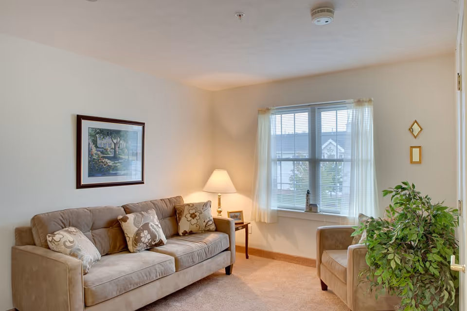 A cozy living room with a beige sofa adorned with patterned cushions, a matching armchair, a side table with a lamp and picture frame, a large window with sheer curtains letting in natural light, a framed painting on the wall, and a green potted plant near the armchair.