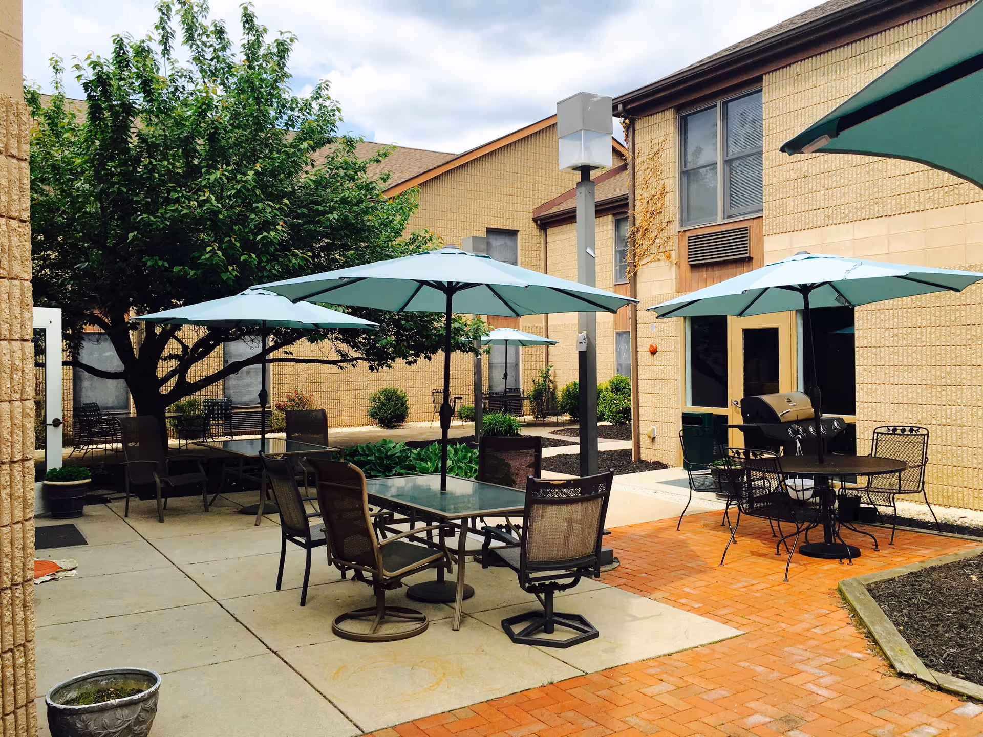 Outdoor patio area with several tables and chairs under light blue umbrellas. There is a barbecue grill near one of the tables. The patio is surrounded by beige brick buildings and some greenery including a tree and bushes.