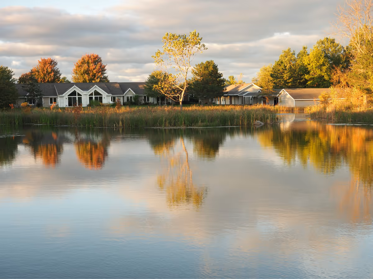 A row of single-story buildings and autumn trees reflected in a calm pond at sunset.