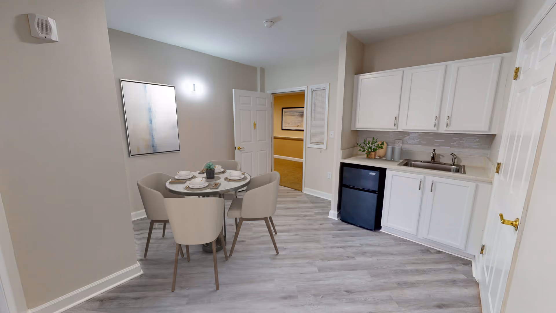 A small dining area with a round table set for four people, surrounded by four beige chairs. To the right is a kitchenette with white cabinets, a small black refrigerator, a sink, and some decorative plants. The floor is light gray wood, and the walls are painted a light beige. A door is open in the background, revealing a hallway with a framed picture on the wall.