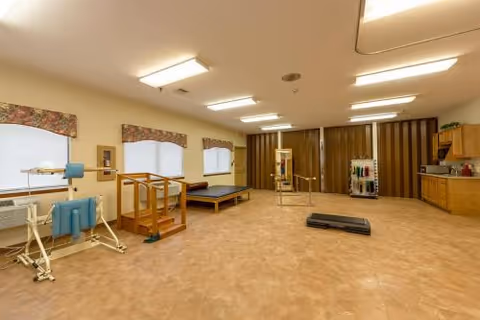 A spacious rehabilitation room with exercise equipment including parallel bars, a treadmill, and padded therapy tables. The room has beige flooring, fluorescent ceiling lights, windows with floral valances, and a kitchenette area with cabinets and a countertop on the right side.