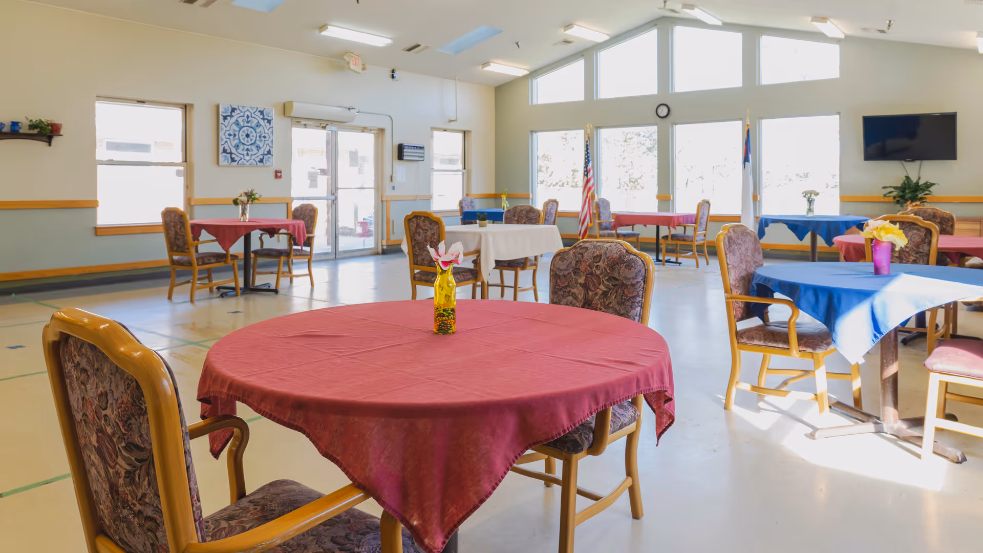 A bright dining room with several round tables covered with red, white, and blue tablecloths. Each table has a small vase with flowers. The room has large windows letting in natural light, a wall-mounted TV, and flags near the windows. Chairs with floral upholstery surround the tables.