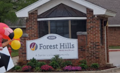 Entrance sign for Forest Hills Main Healthcare, a senior living community, mounted on a brick structure with a small roof. There are some colorful balloons and part of an inflatable flamingo visible on the left side of the image.