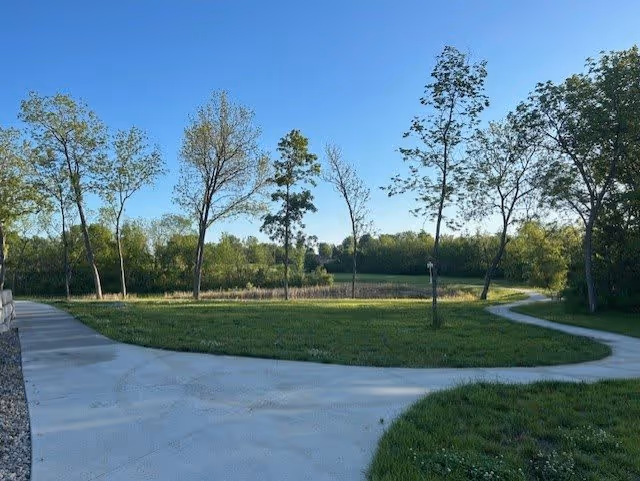 A paved walking path curves through a grassy area with several young trees and a backdrop of dense green foliage under a clear blue sky.
