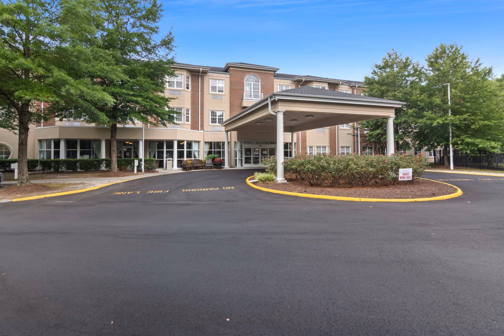 Front entrance of a three-story senior living building with a covered porte-cochere, driveway, and landscaping.
