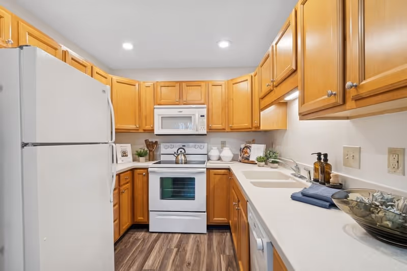 A clean and well-lit kitchen with wooden cabinets, a white refrigerator, a white stove with a kettle on it, a white microwave above the stove, a double sink, and a countertop with decorative items including plants, soap dispensers, and a bowl.