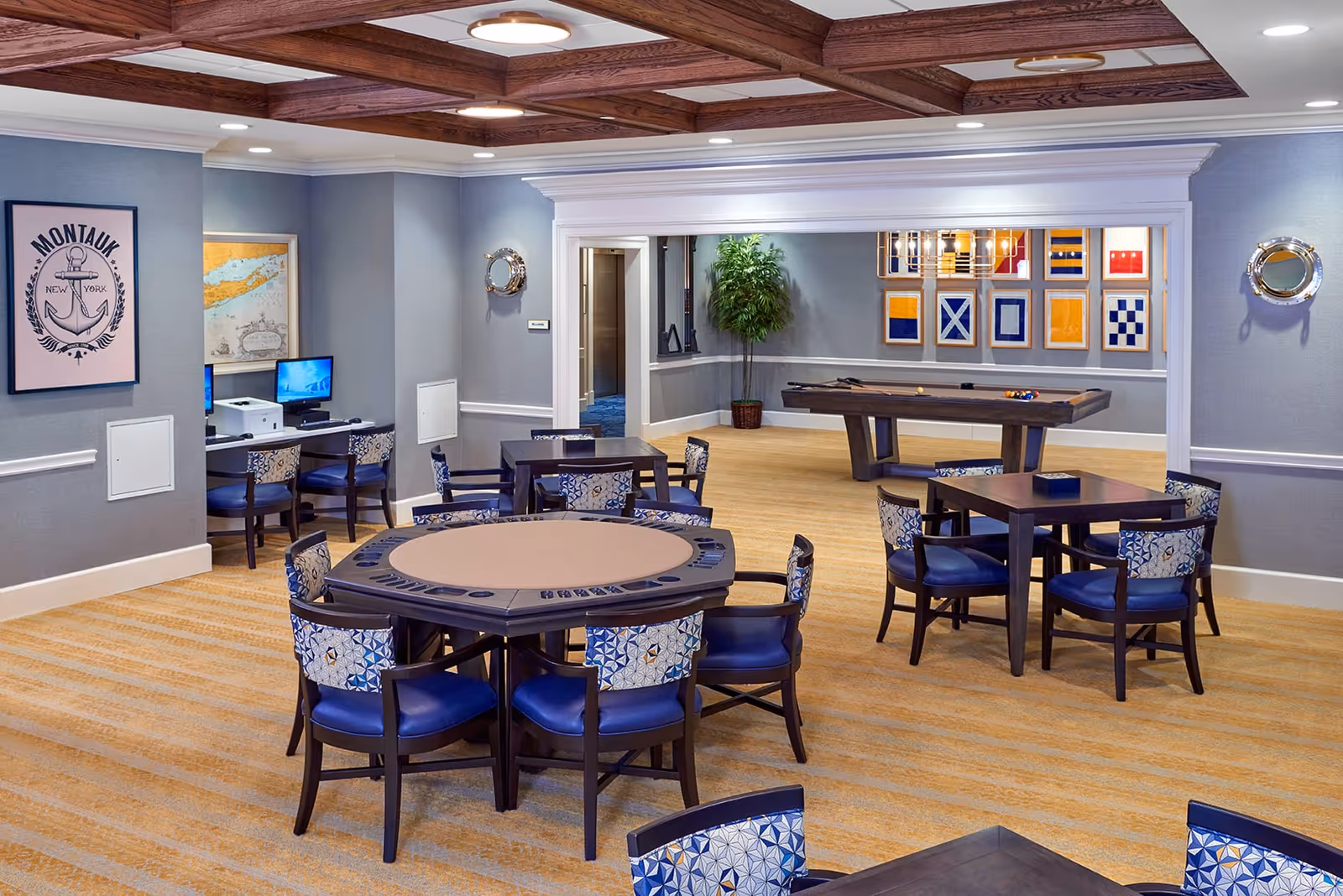A spacious recreational room with multiple tables and chairs, including a poker table and a square table. In the background, there is a pool table with billiard balls set up. The walls are decorated with framed nautical flags and a Montauk New York poster. There are two computer stations with monitors and chairs along one wall, and a potted plant near the pool table. The ceiling has wooden beams and recessed lighting.