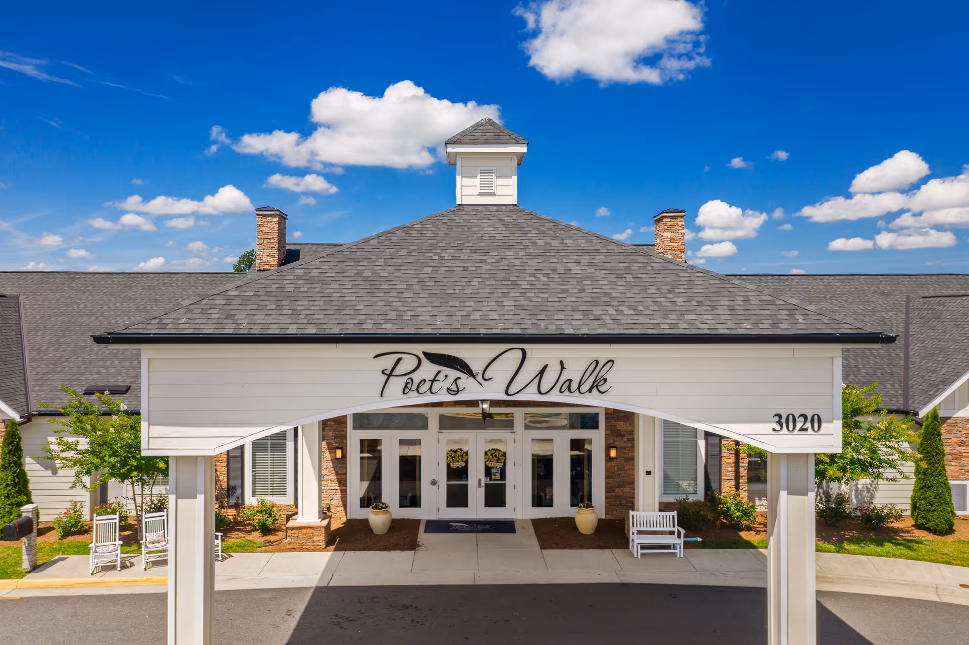 Entrance canopy of Poet's Walk senior living facility showing the covered drive-up, pillars, double glass doors, and benches under a blue sky.