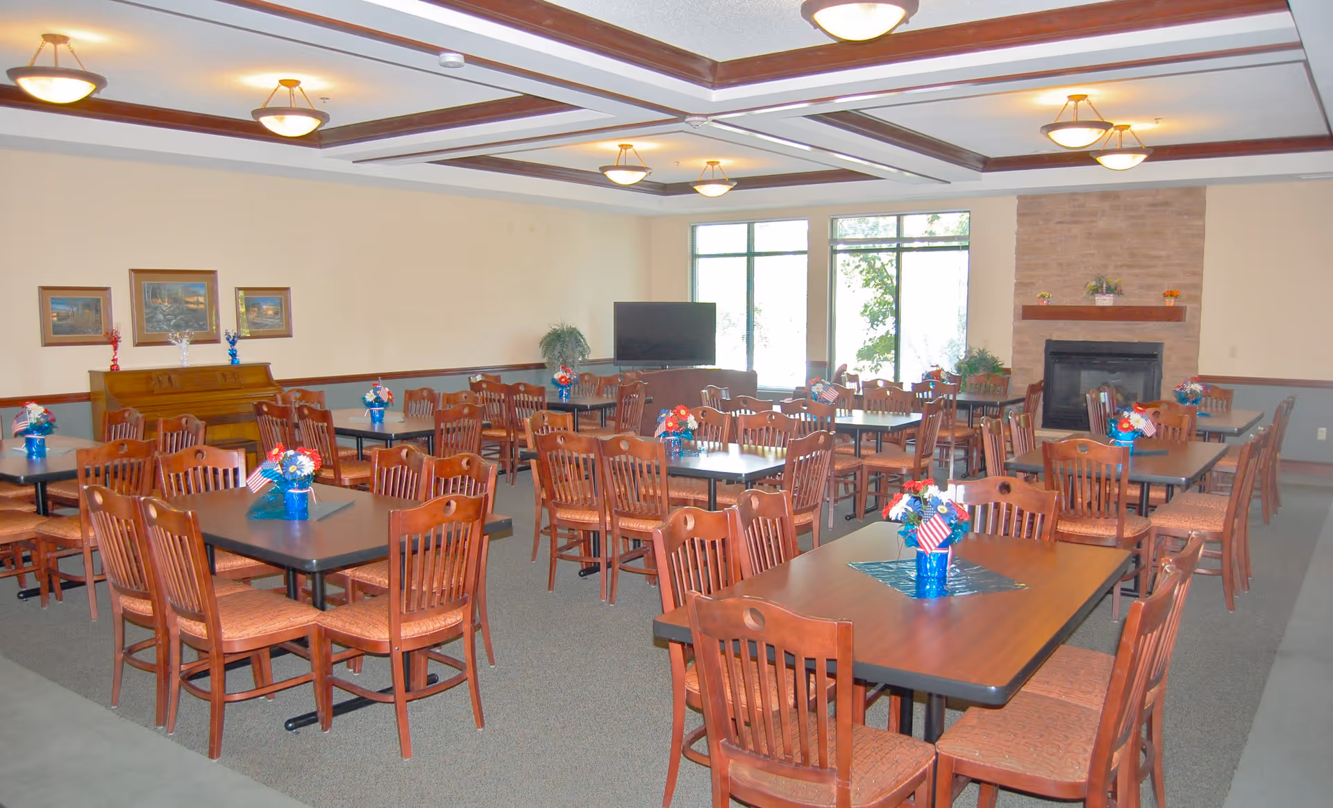 A spacious dining room with multiple wooden tables and chairs arranged neatly. Each table has a small blue vase with red, white, and blue floral decorations and small American flags. The room features a fireplace with a stone surround, a piano against one wall, framed artwork, large windows letting in natural light, and ceiling lights with wooden beams.