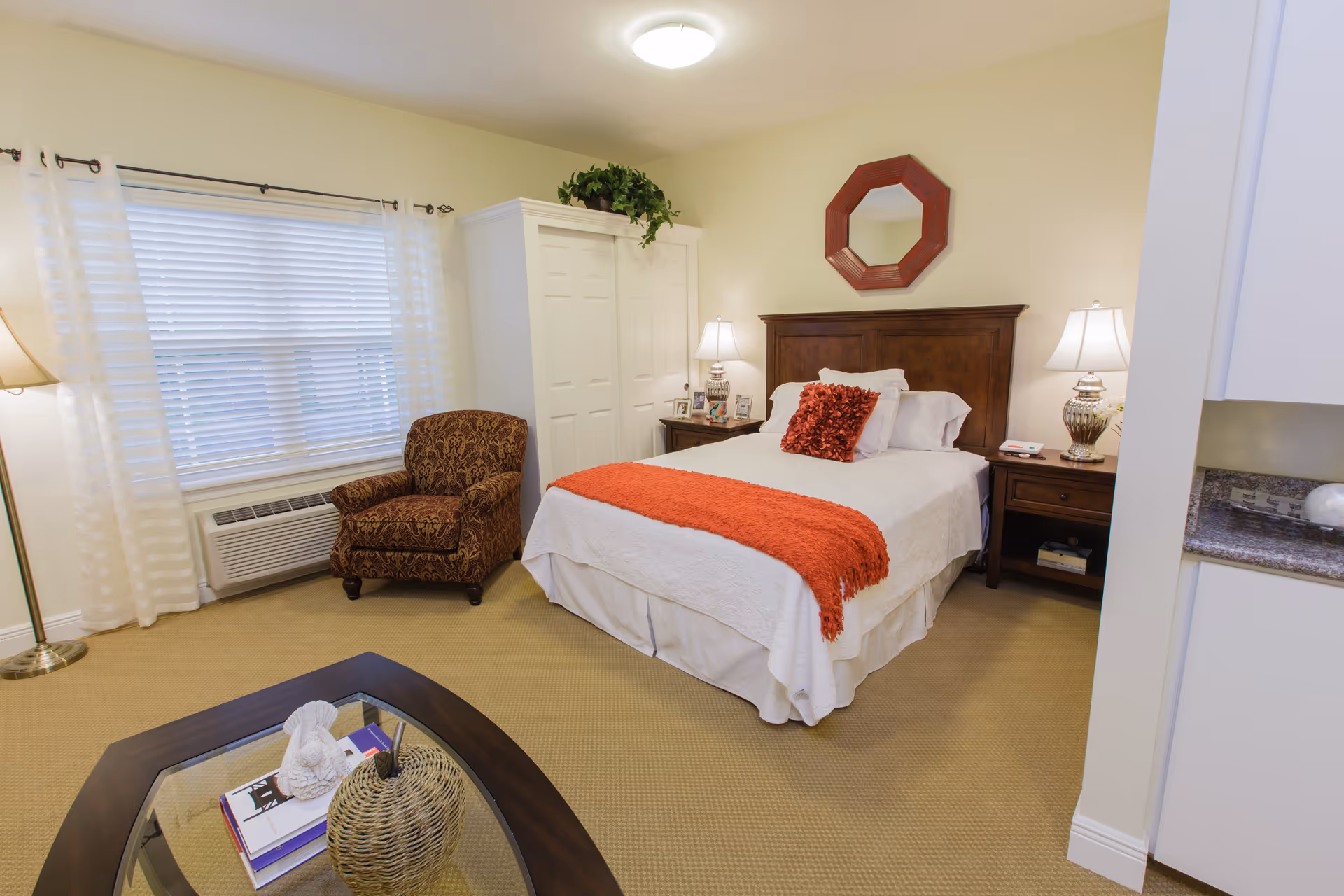 A cozy bedroom with a bed dressed in white linens and an orange throw blanket and pillow. The room features a wooden headboard, two bedside tables with lamps, a patterned armchair near a window with white curtains, a white wardrobe, and a glass coffee table with decorative items on a beige carpeted floor.