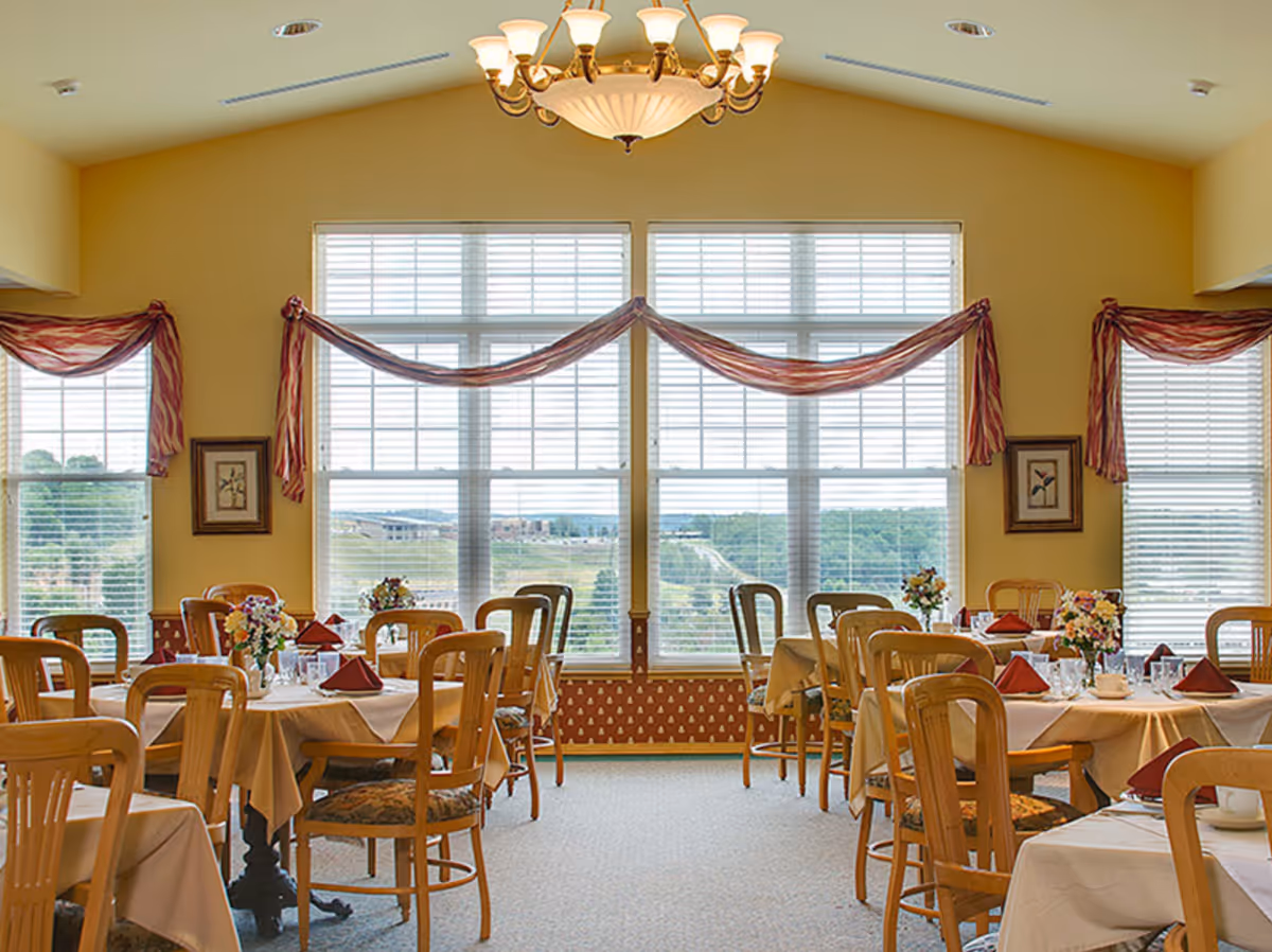 A bright dining room with several tables covered in beige tablecloths, each set with folded red napkins, glassware, and floral centerpieces. Wooden chairs surround the tables. Large windows with white blinds and red swag curtains allow natural light to fill the room. A chandelier hangs from the ceiling.