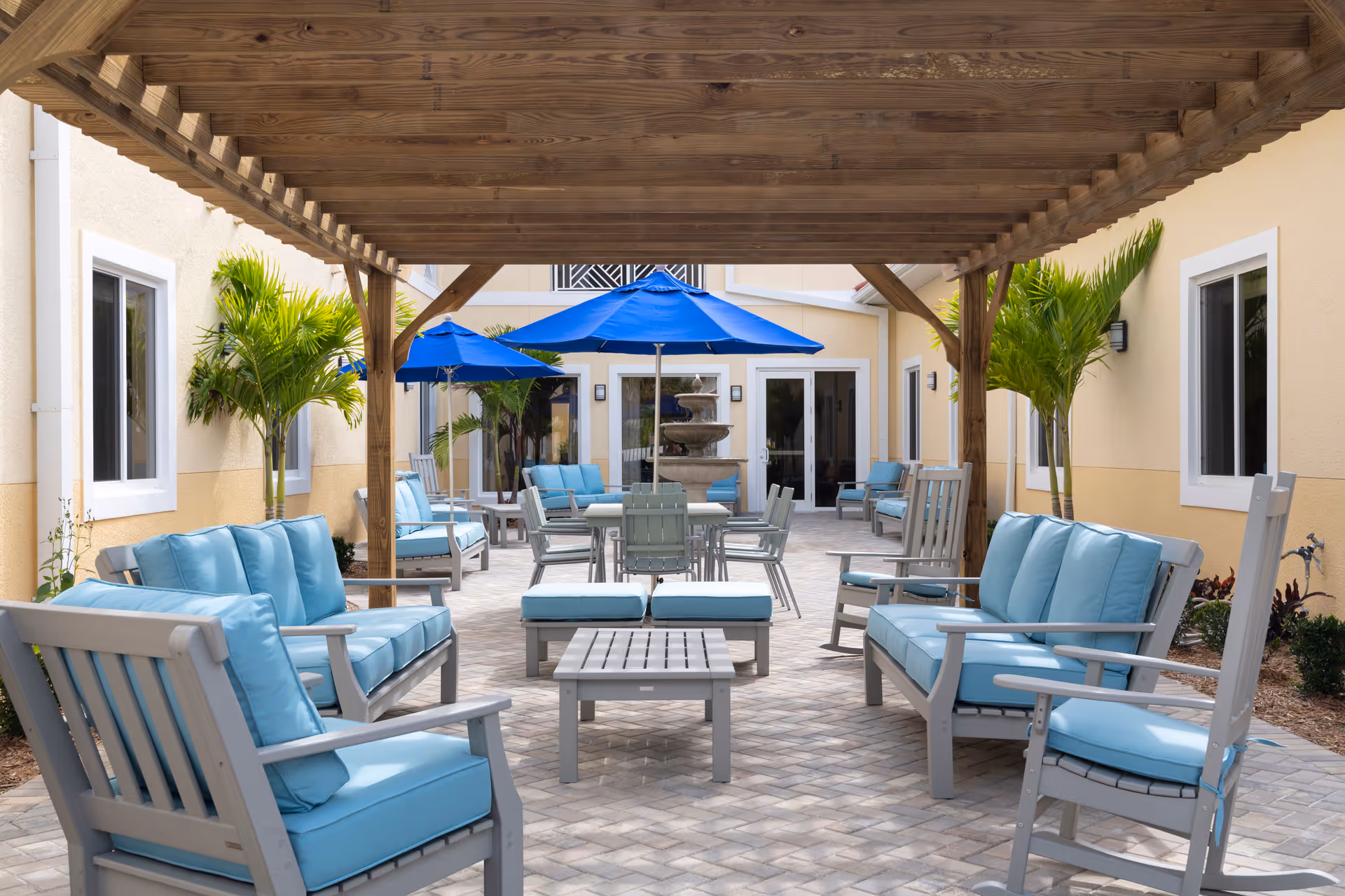 Outdoor seating area at The Pineapple House at Sapphire Lakes featuring light blue cushioned chairs and sofas arranged under a wooden pergola, with blue umbrellas and a multi-tiered water fountain in the background, surrounded by beige building walls and potted palm plants.