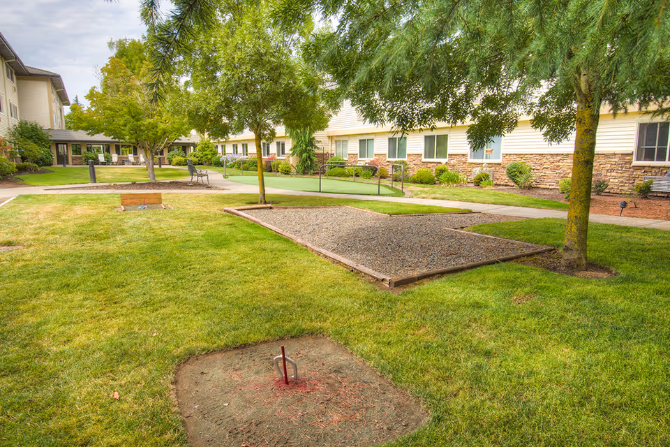 Outdoor area of Cascade Park Retirement Community featuring a grassy lawn, trees, a horseshoe pit, a gravel bocce ball court, benches, and a paved walkway with the building in the background.